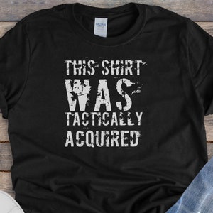 May include: Black t-shirt with white text that reads "This shirt was tactically acquired". The shirt is laid flat on a woodgrain surface with a pair of white sneakers and blue jeans.