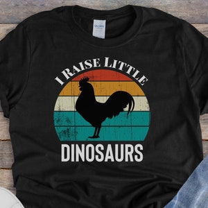 May include: Black t-shirt featuring a vintage-style graphic of a rooster and the text "I raise little dinosaurs".