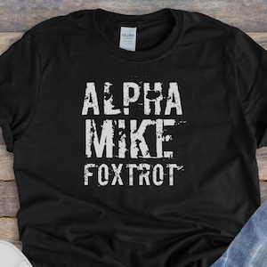 Puede incluir: Camiseta negra con texto blanco que dice "ALPHA MIKE FOXTROT" sobre un fondo de madera. La camiseta está puesta plana con un par de zapatillas blancas y unos vaqueros azules.