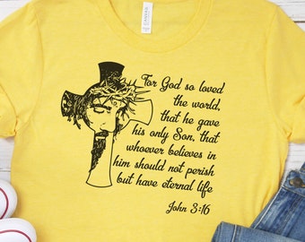 Puede incluir: Camiseta amarilla con una imagen en blanco y negro de una cruz y una cita de la Biblia: "Porque tanto amó Dios al mundo, que dio a su Hijo unigénito, para que todo aquel que en él cree, no se pierda, mas tenga vida eterna. Juan 3:16"