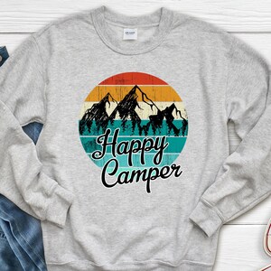 May include: A gray sweatshirt with a retro-style graphic of mountains and trees in a circle. The text "Happy Camper" is printed below the graphic.
