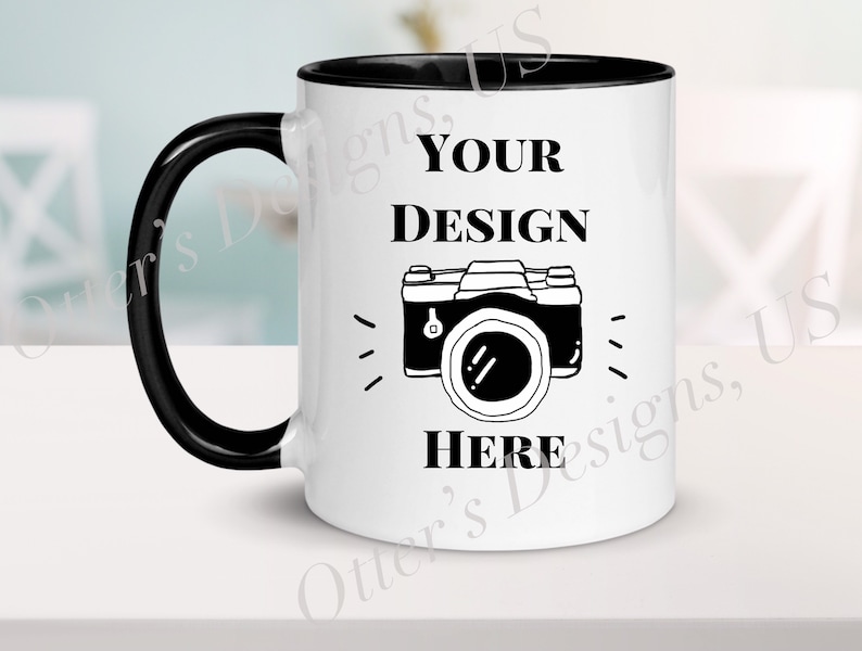 K&ouml;nnte beinhalten: Eine wei&szlig;e Keramiktasse mit schwarzem Henkel und schwarzem Rand. Die Tasse zeigt eine Schwarzwei&szlig;illustration einer Kamera mit dem Text "YOUR DESIGN HERE" darauf.