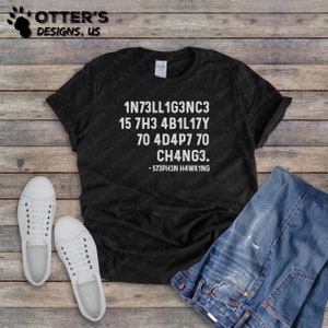 Puede incluir: Camiseta negra con texto blanco que dice "1N73LL1G3NC3 15 7H3 4B1L17Y 70 4D4P7 70 CH4NG3. -573PH3N H4WK1NG". La camiseta está puesta sobre una superficie de madera con un par de zapatillas blancas y unos vaqueros azules.