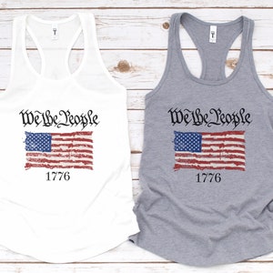 K&ouml;nnte beinhalten: Zwei Tanktops mit einer abgenutzten amerikanischen Flagge und dem Text "We the People" und "1776". Ein Tanktop ist wei&szlig; und das andere ist grau.