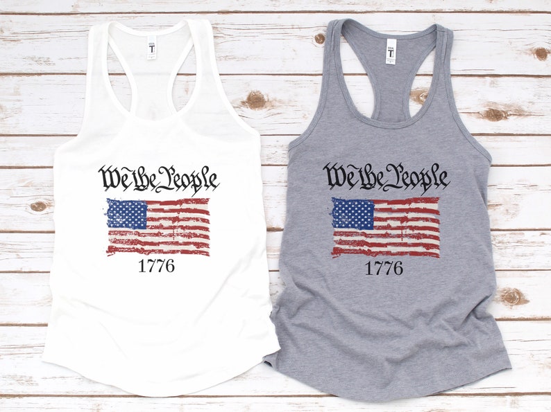 K&ouml;nnte beinhalten: Zwei Tanktops mit einer abgenutzten amerikanischen Flagge und dem Text "We the People" und "1776". Ein Tanktop ist wei&szlig; und das andere ist grau.