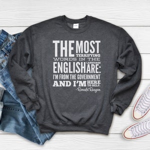 Puede incluir: Una sudadera gris oscura con un estampado gr&aacute;fico blanco que dice "THE MOST TERRIFYING WORDS IN THE ENGLISH ARE: I'M FROM THE GOVERNMENT AND I'M HERE TO HELP. - Ronald Reagan"