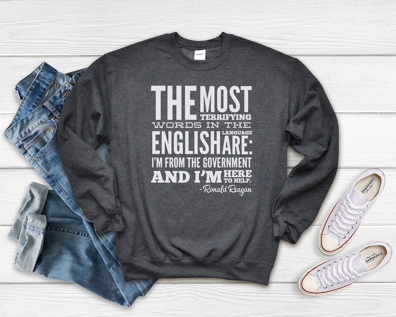 Puede incluir: Una sudadera gris oscura con un estampado gr&aacute;fico blanco que dice "THE MOST TERRIFYING WORDS IN THE ENGLISH ARE: I'M FROM THE GOVERNMENT AND I'M HERE TO HELP. - Ronald Reagan"