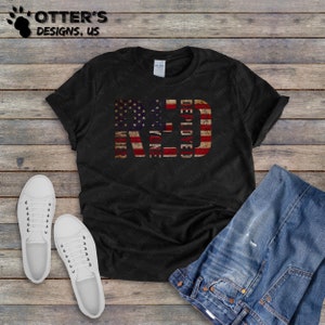 Puede incluir: Camiseta negra con un dise&ntilde;o de bandera americana roja, blanca y azul. Las palabras "RED, WHITE & BLUE" est&aacute;n escritas verticalmente en la bandera, con "DEPLOYED" escrito horizontalmente en la parte inferior.