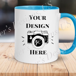 May include: White ceramic mug with a light blue rim and handle. The mug has a black camera graphic and the text "YOUR DESIGN HERE".