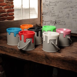 May include: Seven paint can shaped pencil holders in different colors. The holders are made of a gray material with a paint drip design. The colors of the holders are blue, orange, black, green, pink, red, and white.