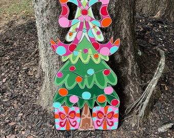 May include: A colorful wooden Christmas tree decoration with a pink and white polka dot bow, green and white lights, and pink and white gift boxes at the base. The tree is hanging from a tree branch with a string of colorful beads.