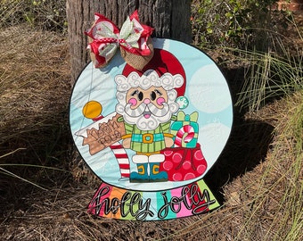 May include: A round, blue and white snow globe door hanger with a cartoon Santa Claus holding a sign that says "North Pole". The snow globe has a red and white striped pole and a red bow with white polka dots on top. The bottom of the snow globe says "holly jolly" in rainbow letters.