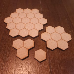 May include: Several unfinished wooden hexagon tiles of varying sizes are arranged on a wooden surface. The tiles are light brown with visible wood grain, and the edges are cleanly cut. The arrangement suggests a game or crafting component.