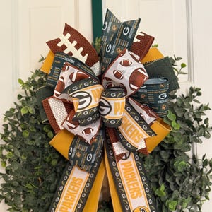 May include: A decorative wreath featuring a large bow with football and Packers-themed ribbons. The bow incorporates green, yellow, and brown ribbons with footballs and the Packers logo. The wreath is set against a backdrop of green foliage.