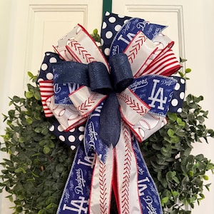 May include: A decorative wreath featuring a large bow with Dodgers baseball team ribbons. The bow has navy blue, white, and red ribbons with baseball and Dodgers logos. The wreath is made of green foliage.