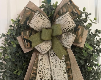May include: A green and brown camouflage ribbon bow with a burlap ribbon with the words "Duty, USA, Defend, Hero, Strong, Brave, Thank you, Service, Army, Selfless, Proud, Freedom" printed on it. The bow is on a green wreath.