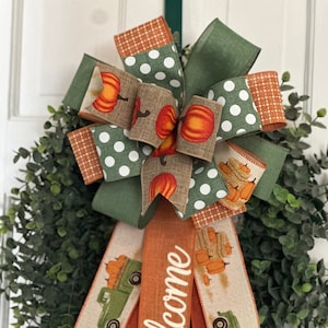 May include: A green, brown, and orange fall wreath with a large bow and the word "Welcome" written on the ribbon. The wreath is decorated with pumpkins, plaid, and polka dots.