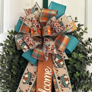 May include: A teal, orange, and brown fall wreath with a welcome sign and a bow. The wreath is decorated with a plaid ribbon, a ribbon with a wheelbarrow and pumpkins design, and a ribbon with a berry and pumpkin design.