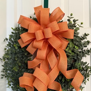 May include: A round green wreath with a large orange bow. The bow is made of wide, textured ribbon and is the focal point of the wreath. The wreath is likely for seasonal or holiday decor.