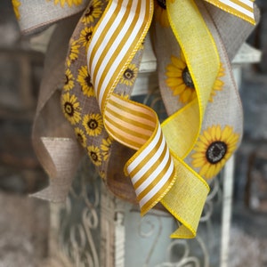K&ouml;nnte beinhalten: Ein gelbes und wei&szlig; gestreiftes Band, das zu einer Schleife gebunden ist, mit einem grob gewebten Band mit Sonnenblumenmuster. Die Schleife ist um eine wei&szlig;e Laterne mit einem dekorativen Metalldesign gebunden.
