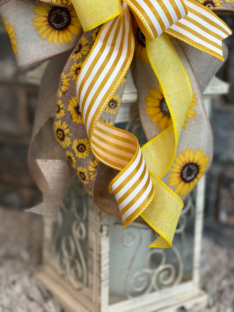 K&ouml;nnte beinhalten: Ein gelbes und wei&szlig; gestreiftes Band, das zu einer Schleife gebunden ist, mit einem grob gewebten Band mit Sonnenblumenmuster. Die Schleife ist um eine wei&szlig;e Laterne mit einem dekorativen Metalldesign gebunden.
