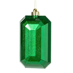 May include: A green, rectangular Christmas ornament with a faceted design, resembling a gemstone. The ornament has a glittery finish and is suspended by a gold-colored metal cap and a thin gold string. The background is white.