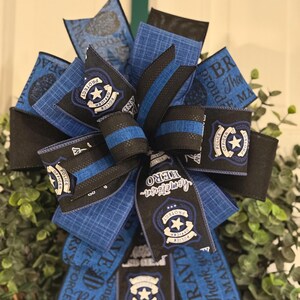 May include: A blue and black wreath with a police officer theme. The wreath is decorated with blue and black ribbons, police badges, and the words "Protect & Serve", "Hometown Hero", "Brave", "Thank you", "Peace Maker", and "Proud".