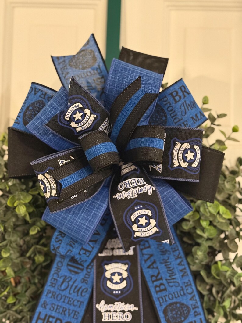 May include: A blue and black wreath with a police officer theme. The wreath is decorated with blue and black ribbons, police badges, and the words "Protect & Serve", "Hometown Hero", "Brave", "Thank you", "Peace Maker", and "Proud".