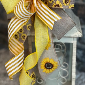 K&ouml;nnte beinhalten: Ein gelbes und wei&szlig; gestreiftes Band, ein einfarbiges gelbes Band und ein Leinenband mit Sonnenblumenmuster sind zu einer Schleife um eine wei&szlig;e Laterne gebunden.