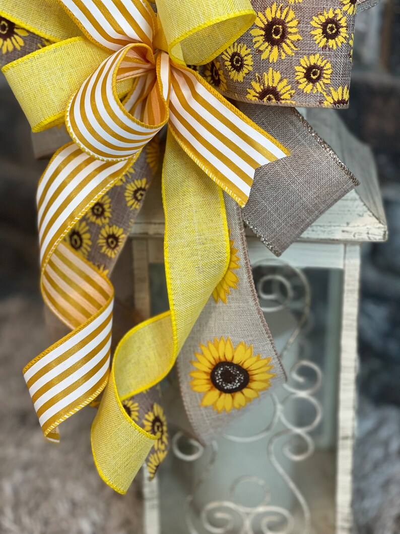K&ouml;nnte beinhalten: Ein gelbes und wei&szlig; gestreiftes Band, ein einfarbiges gelbes Band und ein Leinenband mit Sonnenblumenmuster sind zu einer Schleife um eine wei&szlig;e Laterne gebunden.