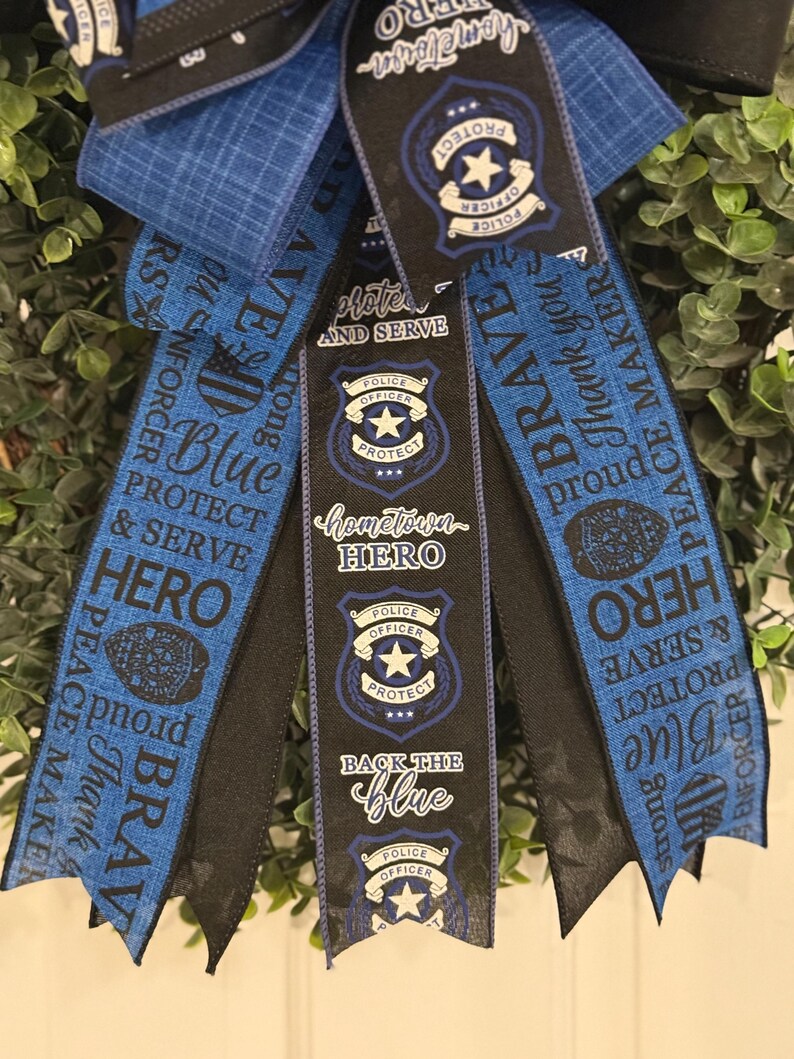 May include: Blue and black ribbon wreath with the words "Protect and Serve", "Hometown Hero", "Back the Blue", and "Brave" printed on them. The ribbons also feature a police officer badge with a star.