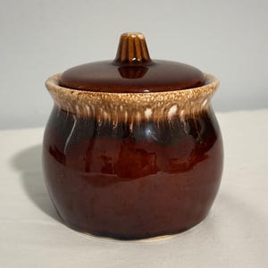 May include: A brown ceramic sugar bowl with a lid. The bowl has a white glaze on the top edge and a brown glaze on the bottom. The lid has a knob on top.