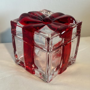 May include: A clear glass gift box with a red bow on top. The box is square-shaped with a lid and is decorated with red ribbon accents. The bow is a deep red color and adds a festive touch to the box.