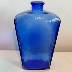 May include: A cobalt blue glass bottle with a textured surface. The bottle has a square base that tapers upwards to a rounded neck. The bottle is empty and sits on a white surface.