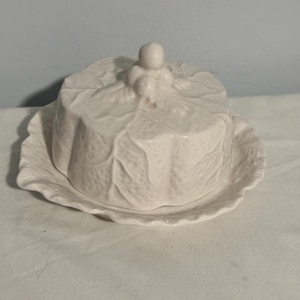 May include: White ceramic butter dish with a lid and base. The lid is shaped like a cabbage with a small decorative figure on top. The base is a scalloped plate. The dish is designed to hold butter and keep it fresh.