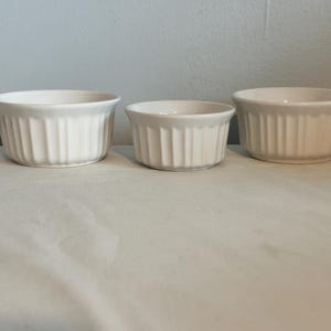 May include: Three white ceramic ramekins with vertical ribbed sides. The ramekins are of varying sizes, perfect for individual servings of desserts or side dishes. They are arranged on a white surface.