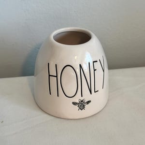May include: A small, off-white ceramic honey pot with a wide opening. The word "HONEY" is printed in black, bold letters on the front, with a small bee illustration below. The pot has a rounded shape.