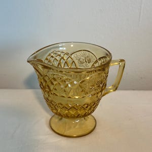 May include: Amber-colored glass creamer with a diamond pattern and floral accents. The creamer has a handle and a spout for easy pouring. It sits on a small, round base. A vintage-style piece for serving cream or other liquids.