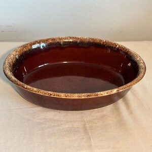 May include: An oval-shaped ceramic baking dish with a deep brown, glossy interior and a textured, mottled rim featuring shades of cream, tan, and dark brown. The dish has a smooth, reflective surface and a wide, shallow basin for food preparation.