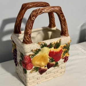 May include: A ceramic basket-shaped container with a woven design and two brown handles. The basket is decorated with colorful fruit, including grapes, apples, pears, and berries. The container is off-white.