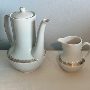 May include: A white ceramic coffee pot with a matching creamer. Both pieces have a gold-colored decorative band around the base. The coffee pot has a lid and a curved handle. The creamer has a handle and a spout.