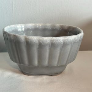 May include: A gray ceramic bowl with a fluted design. The bowl has a rectangular base and a slightly flared rim with a white, dripped glaze. The bowl is suitable for serving or as a decorative piece.