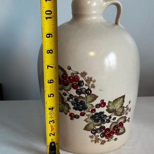 May include: A beige ceramic jug with a handle, decorated with a floral pattern of red and black berries and green leaves. The jug is approximately 11 inches tall.