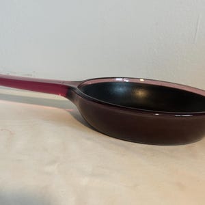 May include: A dark purple glass serving dish with a long handle. The dish has a deep bowl and a smooth, glossy finish. The handle is the same color as the bowl. The interior of the bowl is black.
