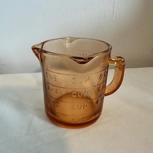 May include: A vintage amber glass measuring cup with a handle and a pouring spout. The cup has measurement markings in cups, including 1/2, 3/4, and 1 cup. The cup is made of transparent glass.