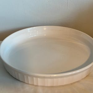 May include: A white, round ceramic baking dish with a fluted exterior. The dish has a shallow depth and a smooth, glossy interior. Ideal for baking casseroles, pies, or other dishes. The dish is sitting on a white surface.