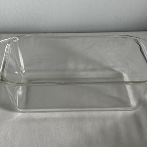 May include: A clear glass loaf pan with a rectangular shape and slightly rounded edges. The pan has a smooth surface and is designed for baking bread.