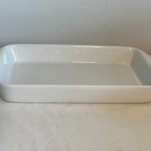 May include: A rectangular, white ceramic baking dish with rounded corners and a slightly raised rim. The dish is empty and sits on a white surface. The overall shape is a long rectangle, suitable for casseroles or baked goods.