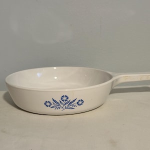 May include: White ceramic skillet with a single handle. The skillet features a blue floral design on the side. The handle is integrated into the design. The skillet is likely used for cooking and serving food.
