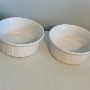 May include: Two white, oval-shaped ceramic ramekins. Each has a ribbed exterior and a smooth, glossy interior. The ramekins are empty and sit on a white surface, suitable for baking or serving individual portions.